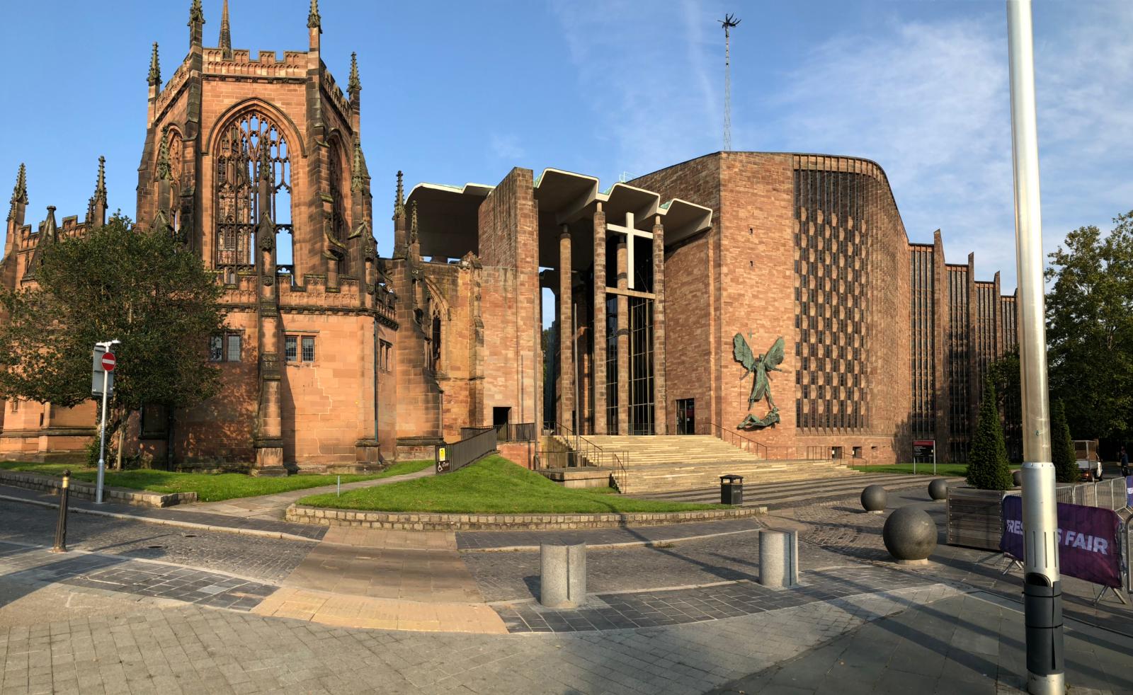 a large stone building with a clock on the side of a road with Coventry Cathedral in the background